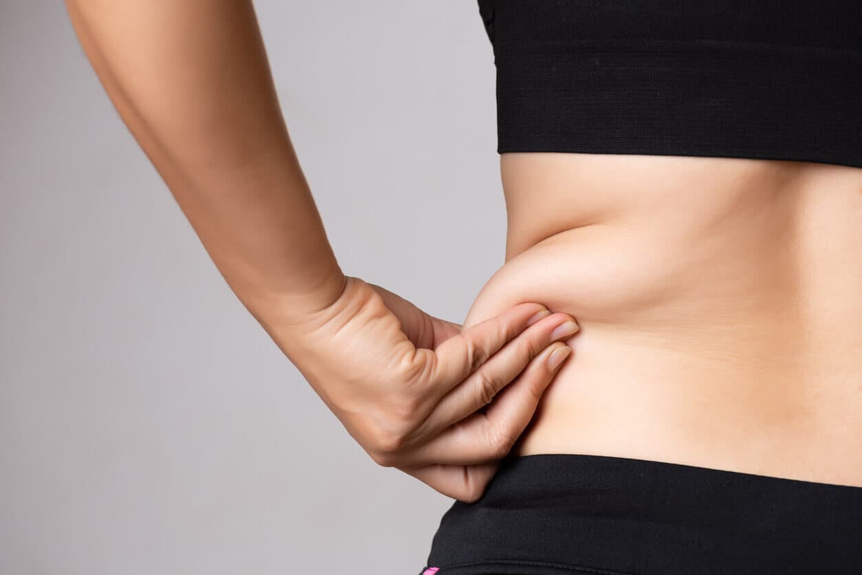 Fundo cinza com apenas o torso de uma paciente visível a frente, segurando com uma mão o excesso de gordura na região das costas e do flanco