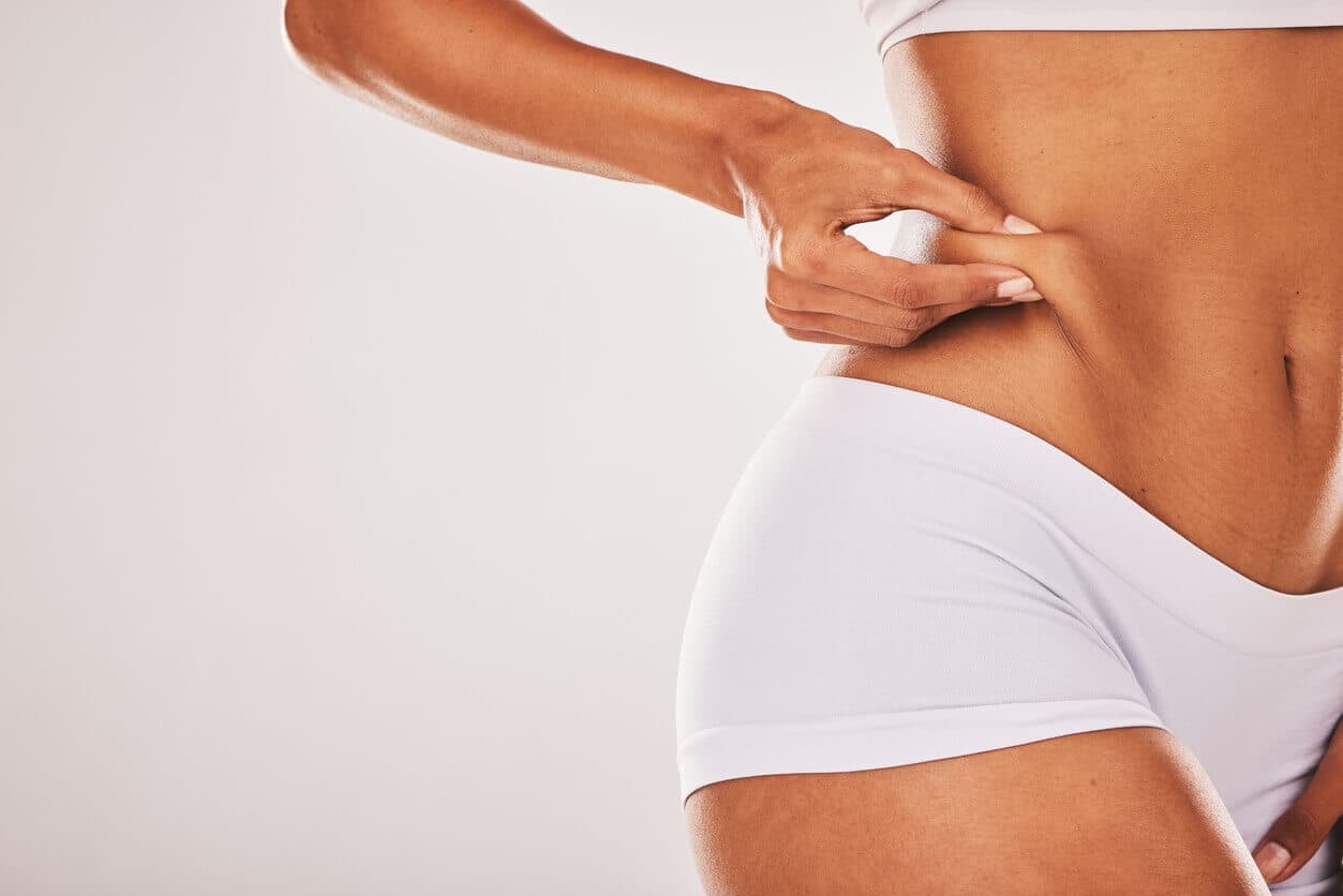 Mulher em roupa íntima branca apertando a lateral do abdômen, destacando a região abdominal para ilustrar procedimentos como abdominoplastia e contorno corporal.