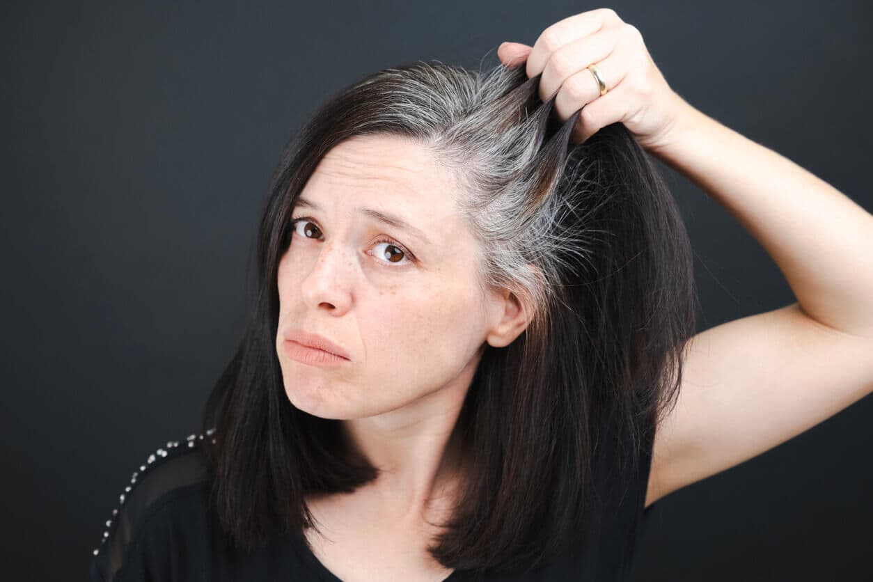 Mulher com expressão triste e insatisfeita enquanto segura mechas do cabelo, revelando raízes grisalhas e enfraquecidas devido o processo de envelhecimento capilar