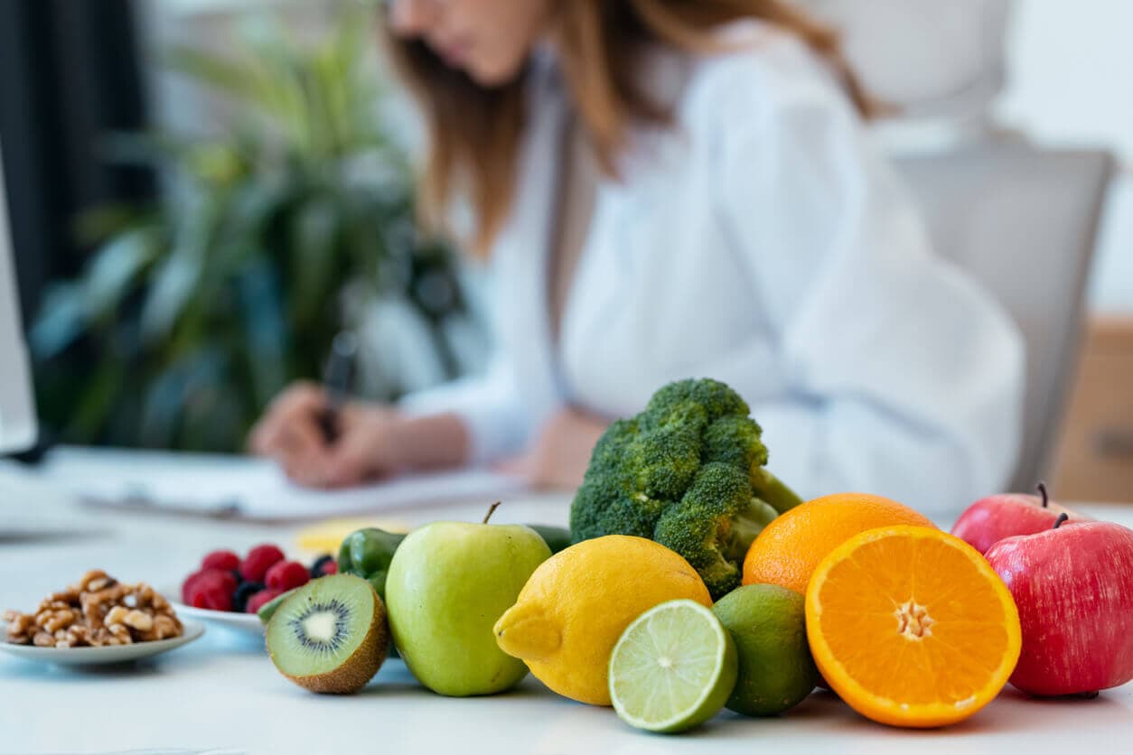 Médica nutricionista, sentada e trabalhando ao fundo, com frutas e legumes arranjados na mesa a frente.
