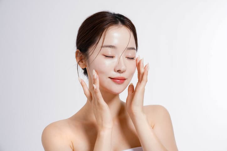 Mulher jovem coreana, tocando as mãos no rosto, com uma pele viçosa e brilhosa, exemplo de uma glass skin