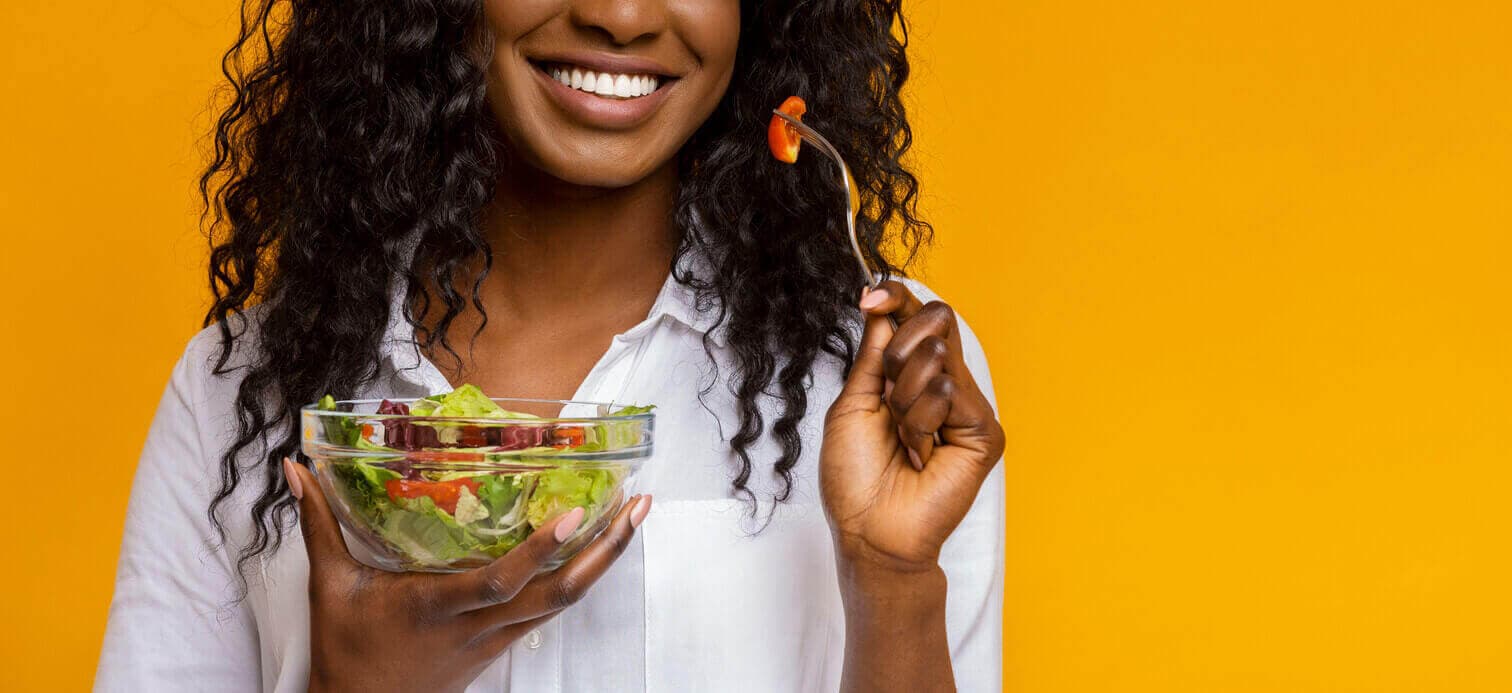 Mulher sorridente segurando uma tigela de salada e comendo com um garfo, representando alimentação leve e saudável para recuperação.