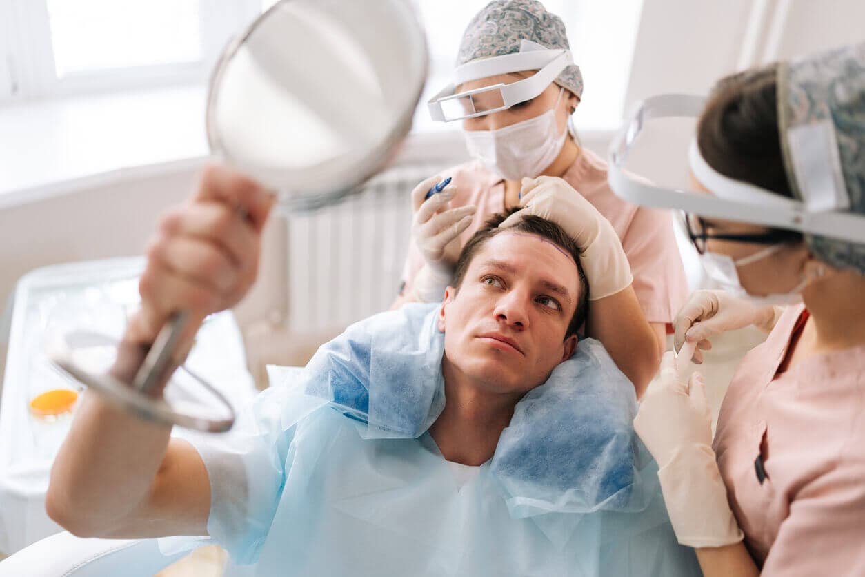 Paciente do sexo masculino segurando um espelho em uma das mãos, enquanto duas médicas cirurgiãs fazem preparações para a realização de um transplante capilar
