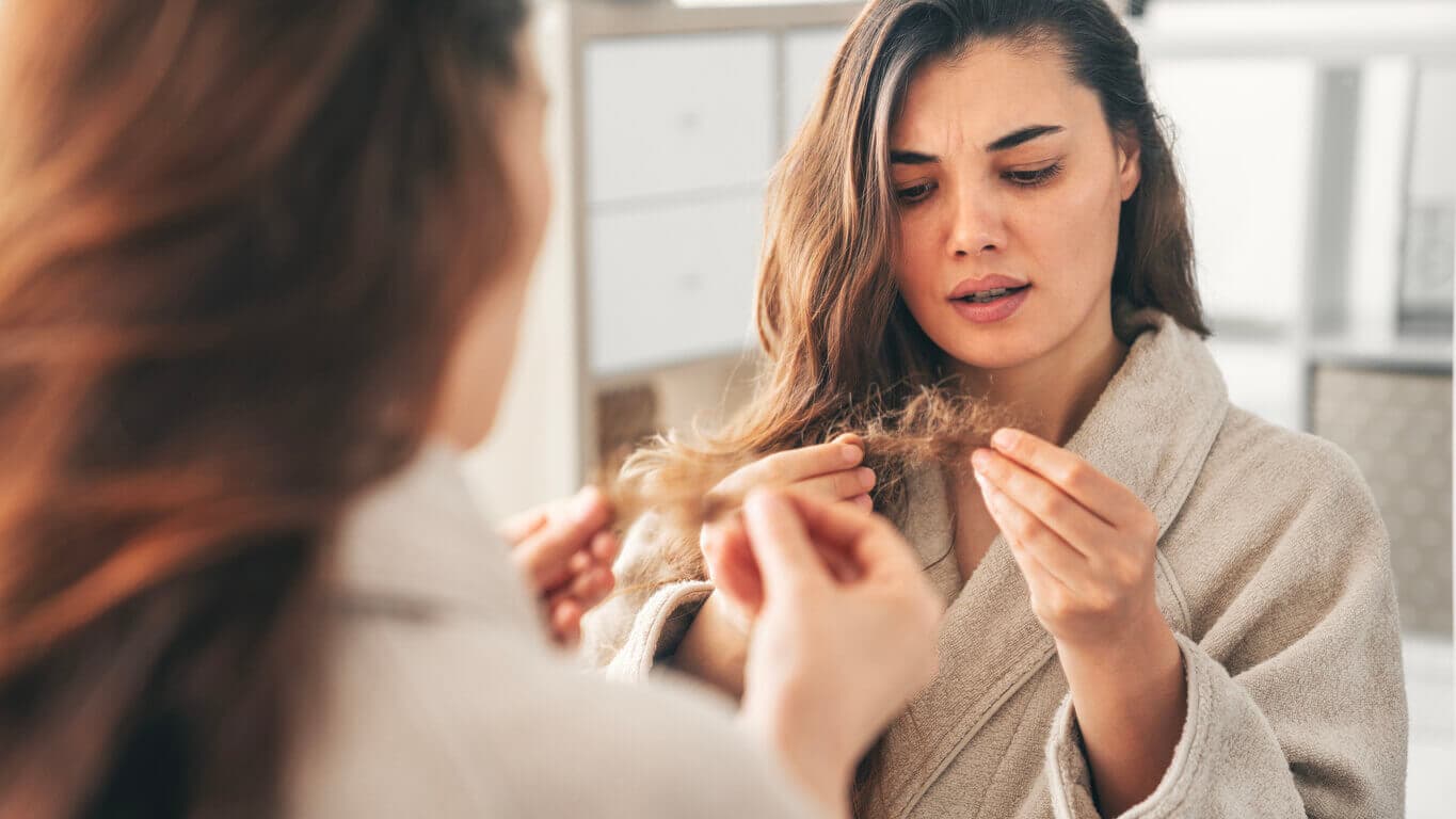 Mulher jovem, se olhando no espelho, com um tufo de cabelo nas mãos, percebendo que apresenta quadro de queda de cabelo