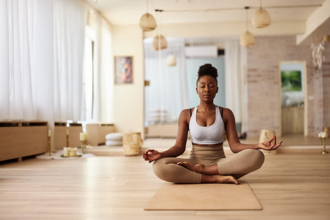 Mulher sentada em posição de lótus sobre um tapete de yoga, meditando em um ambiente claro e tranquilo de estúdio, com olhos fechados e expressão serena