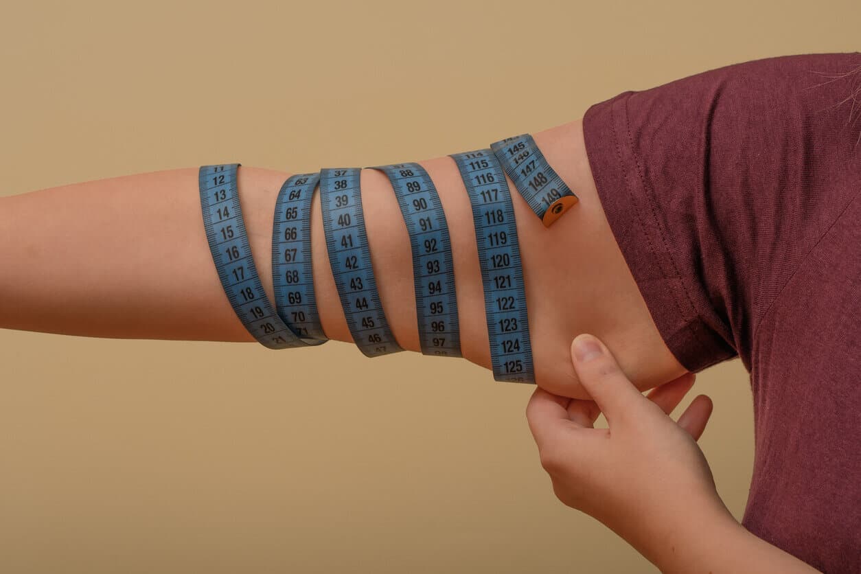 Close de um braço, enrolado em fita métrica, com uma mão segurando o excesso de flacidez corporal